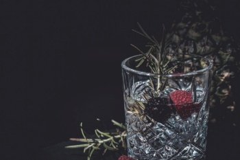 Gin-tonic alcoholic cocktail. Berries, rosemary, ice. On a black wooden background.