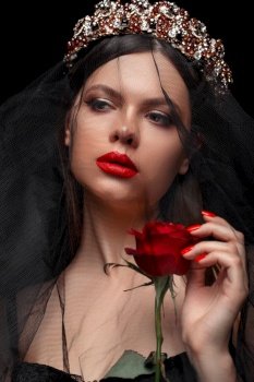 young beautiful girl in a black headband and black veil in a corset with a scarlet rose in her hands on a black isolated background.