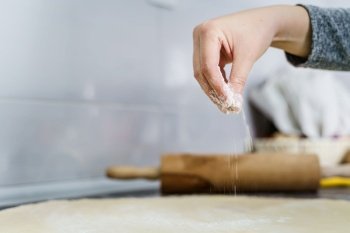 Close up on hand of unknown woman holding flour and spreading over prepared dough on the kitchen counter making homemade paste or croissants at home