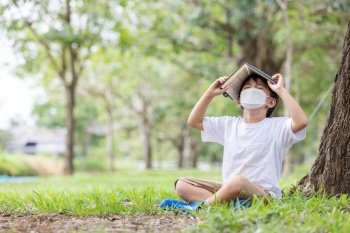 A white boy wearing a hygienic mask sits alone under a tree reading a book covering his head.