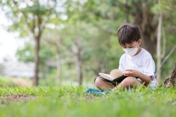 A white boy wearing a hygienic mask sits alone under a tree reading a book.
