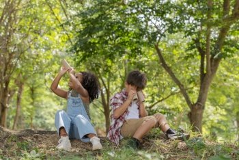 Multi-ethnic children in casual clothing sitting on tree roots, looking at through tissue tube paper to used instead binocular .Exploring nature and camping summer concept.