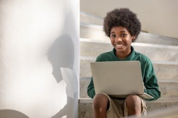 An African curly haired boy sits on a laptop in the corridor downstairs under a building