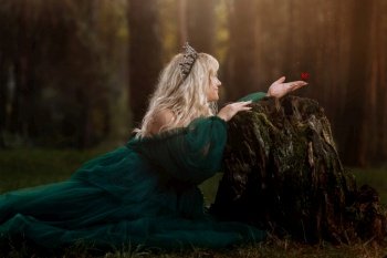 A beautiful blonde young woman in a long green dress and a diadem on her head in the forest. girl sitting near the old stump with butterfly on her hand. Solar glare. Fantasy. fairy tale.. A beautiful blonde young woman in a long green dress and a diadem on her head in the forest. girl sitting near the old stump with butterfly on her hand. Solar glare. Fantasy. fairy tale