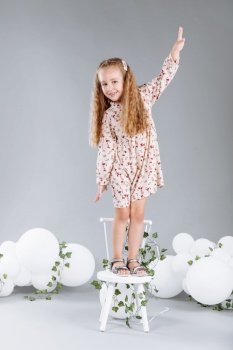 Cute little girl blonde smiling having fun in studio with white balloons and green. child in floral dress standing on white chair and celebration easter. happy childhood.. Cute little girl blonde smiling having fun in studio with white balloons and green. child in floral dress standing on white chair and celebration easter. happy childhood