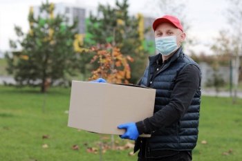 Delivery man in red cap white t-shirt uniform face medical mask gloves hold empty cardboard box on yellow studio background. Service coronavirus. Online shopping. mock up.. Delivery man in red cap, face medical mask, gloves hold empty cardboard box outdoors. Service coronavirus. Online shopping. mock up.