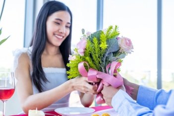 Valentine’s day and asian Young happy couple concept, a man holding a bouquet roses Give to woman with hands over smiling her face awaits surprise after lunch In a restaurant background