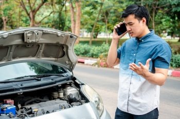Angry Asian man and using mobile phone calling for assistance after a car breakdown on street. Concept of vehicle engine problem or accident and emergency help from Professional mechanic