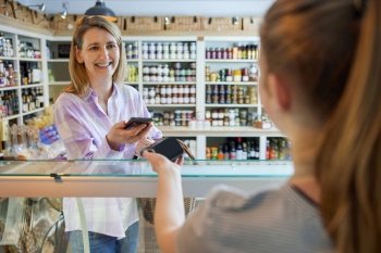 Smiling Female Customer Delicatessen Food Store Making Contactless Payment With Mobile Phone For Shopping To Sales Assistant