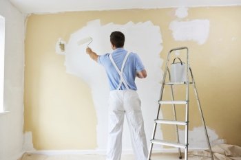 Rear View Of Man Wearing Overalls Painting Wall In Room Of House With Paint Roller
