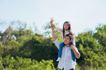 Happy Asian young family father and carrying an excited girl on shoulders having fun and enjoying outdoor lifestyle together playing aircraft toy on sunny summer day, Father’s day concept