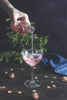 Woman’s hand holds a bottle and pouring champagne in elegant wineglass on dark wooden table surface with dry roses, close up, shallow depth of the field