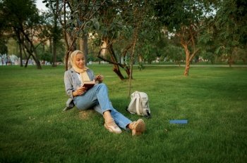 Arab female student in hijab reading textbook in summer park. Muslim woman with books resting on the lawn. Religion and education. Arab female student reading textbook in park