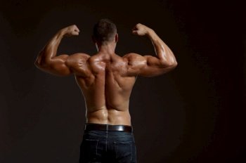 Male muscular athlete poses in studio, back view, dark background. One man with athletic build, shirtless sportsman in jeans pants, active healthy lifestyle. Male muscular athlete poses in studio, back view