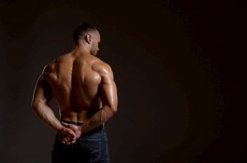 Strong male athlete poses in studio, back view, dark background. One man with athletic build, shirtless sportsman in jeans pants, active healthy lifestyle. Strong male athlete poses in studio, back view