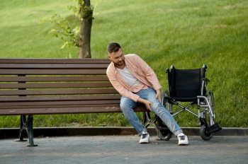 Disabled man transfers from a wheelchair to a bench in the park. Paralyzed people and disability difficulties, handicap overcoming. Handicapped male person walking along the alley. Disabled man transfers from wheelchair to a bench