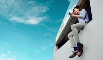 Happy Smiling Young Businessman in Casual wear Using Mobile Phone at the high rooftop Urban City Sky. Low Angle View