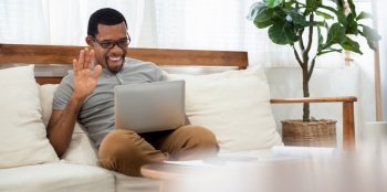 Bearded African American man in eyeglasses sitting on sofa making online video call on laptop. Smiling Black male enjoy with internet technology while talking with friends and family on notebook 