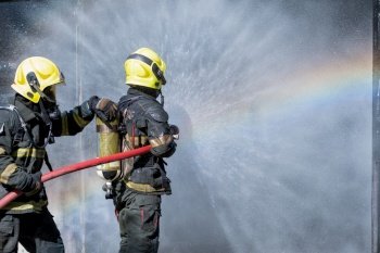 Two fireman or firefighters stand and help each other to spray of water with curtain shape and rainbow reflex occur on water aerosol in front of container containing fire inside.