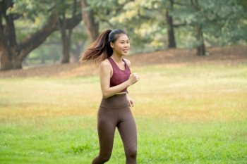 Wide shot of Asian woman jogging in park or garden with green field and tree as background during morning time.