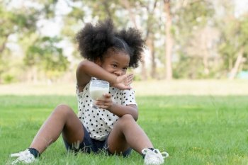 African little kid girl wiping milk stain on her lips with her hand. Cute child holding a glass of milk and sitting on green grass at park on summer