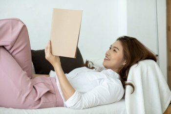 Beautiful Attractive Asian woman lying on sofa in living room reading book for learn and education smile and positive thinking feeling so happiness and comfortable,Education Concept