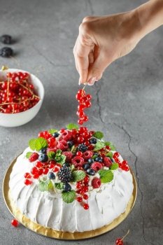 Pavlova cake with cream and fresh summer berries. Close up of Pavlova dessert with forest fruit and mint. Food photography. The process of decorating the cake with red currant berries. Hands on the photo