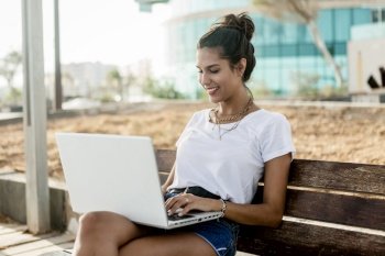 Happy young woman in summer outfit smiling and browsing social media on modern laptop while sitting on wooden bench on street. Smiling woman using laptop on bench