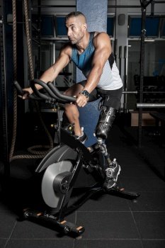 Confident athletic man with artificial leg limb riding stationary bicycle in gym. Sportive man with prosthesis leg riding bicycle