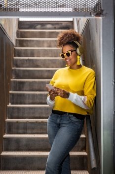 Happy youthful African American female in trendy yellow outfit and sunglasses listening to music through wireless headphones and messaging on smartphone while standing on staircase. Trendy hipster black woman in headphones browsing smartphone