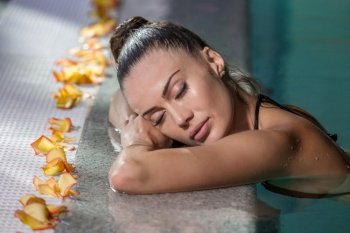 Side view sensual female with closed eyes leaning on poolside near delicate petals while enjoying spa procedure in pool water. Woman with closed eyes in swimming pool