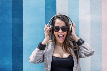 Young female in smart casual outfit and sunglasses touching headphones and yelling in excitement while listening to music against striped wall. Excited woman listening to music and screaming