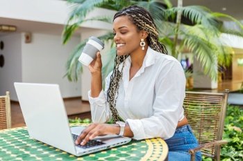 Cheerful African American woman using laptop and holding cup of coffee outside. Woman with braids sitting using laptop and drinking coffee.. Cheerful African American woman using laptop and holding cup of coffee outside