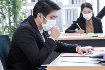 A young Asian business man is wearing mask and coughing while working in the office with blur background of colleague