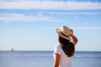 Beautiful girl looks at the sea. Young girl in a hat looking at a calm sea and blue skies back view.