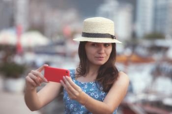Young travelling woman in hat taking selfie photos over summer sunny mediterranean city with smartphone camera. Brunette girl making photography on summer vacation. Young travelling woman in hat taking selfie photos over summer sunny mediterranean city with smartphone camera.