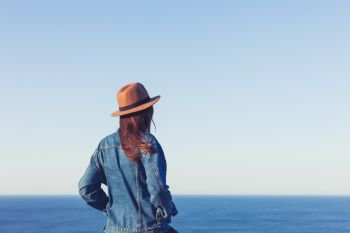 Closeup back view of woman in jeans jacket and hat standing and looking at blue ocean and sky.