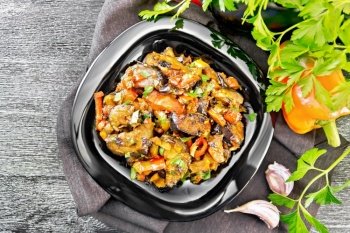 Vegetable ragout with eggplant, tomatoes, sweet and hot peppers, onions, carrots, fried with herbs and spices in a plate on towel, garlic, parsley on dark wooden board background from above