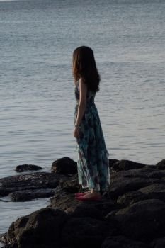 A young thin teenage girl in a long dress, standing on a rocky coast, looking far in the sea. We see her from the back. Assol