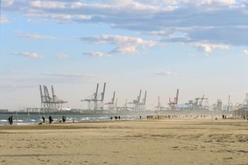 People walking on the beach of Valencia in winter, Spain. Scene with industrial port and container cranes. Valencia beach and industrial port, Spain