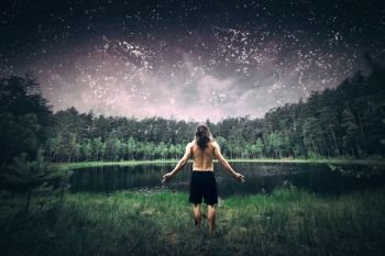 Young man standing in the forest with arms wide open. Inspirational view of a summer night sky full of stars, lake and woods. Nature and freedom concept.. Young man standing in the forest at night.