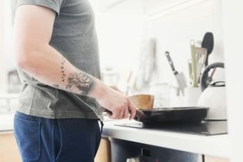 Mid-body shot of a man standing in front of the stove, preparing the meal on a frying pan.. Man frying a meal in the morning