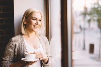 Young woman standing by the window with a cup of coffee, looking outside and smiling. Natural outdoor light.. Woman with a cup of coffee standing by the window.