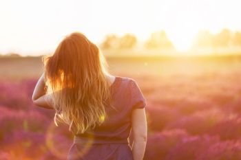 Young woman touching her long sombre hair looking at lavender field at sunset. Romantic warm sunshine. Young woman touching her long sombre hair looking at lavender field at sunset
