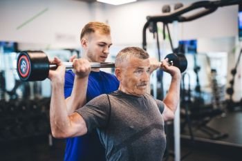 Senior man lifting weights with help of gym assistant. Work of personal trainer. Bodybuilding.. Senior man lifting weights with help of gym assistant.