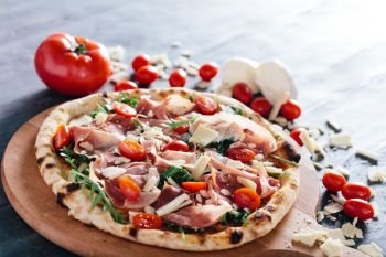 Pizza with ham, cheese, tomatoes and rocket salad on wooden board. Tomatoes, mozzarella and parmesan in the background. Popular traditional food.. Pizza with ham, cheese, tomatoes and rocket salad .