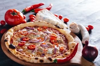 Pizza with hot pepper, red onion, sausage and blue cheese on wooden board. Tomatoes, red onion, blue cheese, sausage and hot pepper in the background. Popular traditional food.. Pizza with hot pepper, red onion, sausage and blue cheese