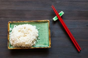 top view of portion of boiled rice on green plate and red chopsticks on rest on dark brown table