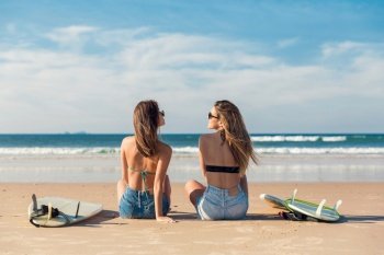 Two beautiful female friends at the beach sitting on the sand close to her surfboards while looking to the ocean