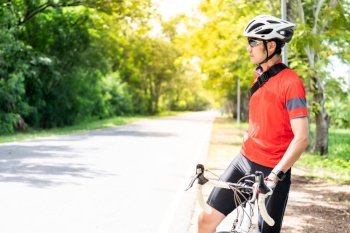 Portrait of asian male cyclist in sportsware shirt with his bicycle in countryside road with green tree in background. Weekend outdoor sport athlete and fitness concept.